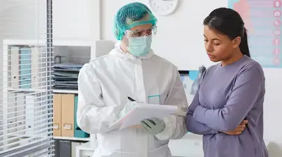 A medical professional wearing PPE speaks with a pensive looking patient.